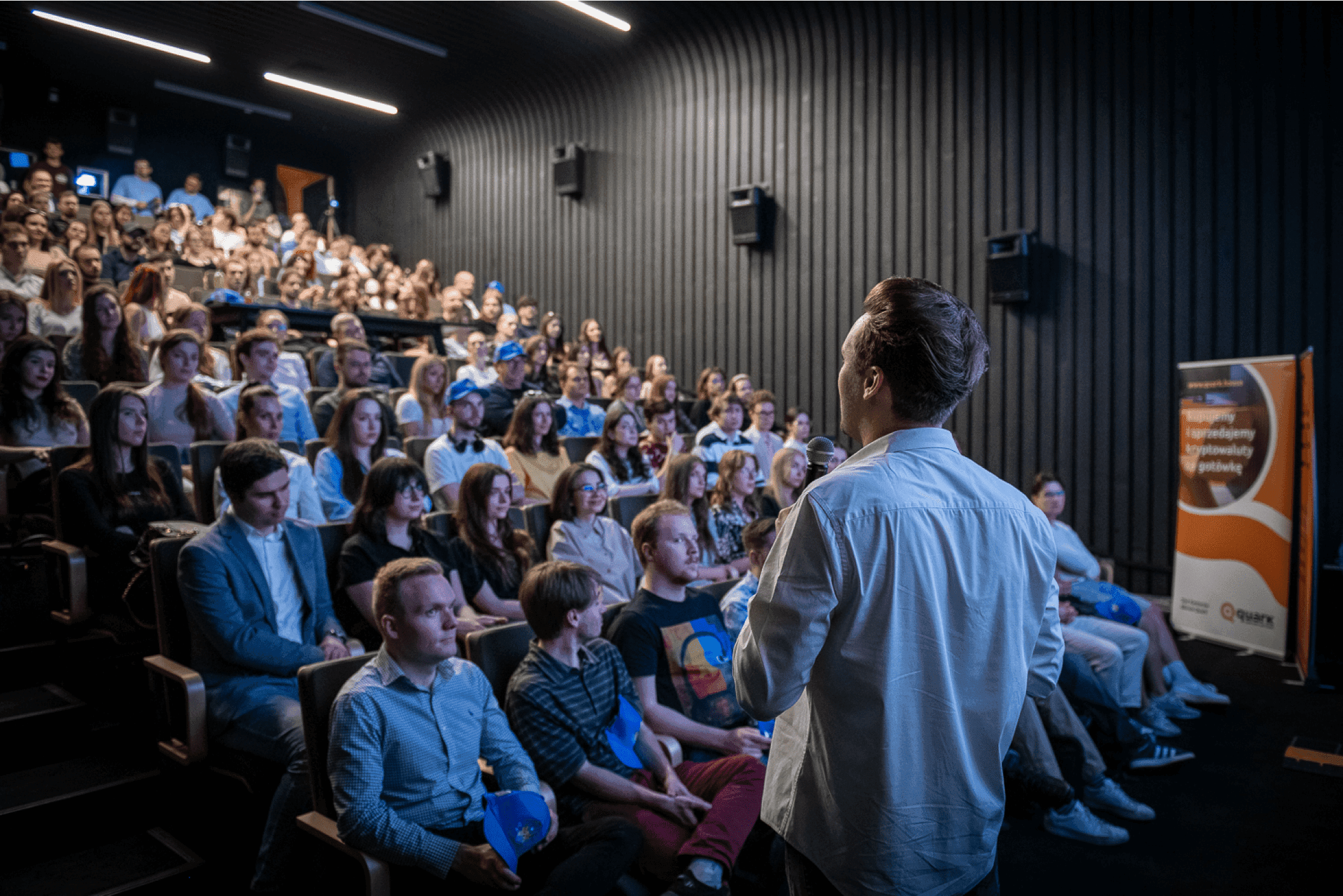 Speaker presenting to a full audience in a dark auditorium, with attendees attentively listening and engaged in the presentation.